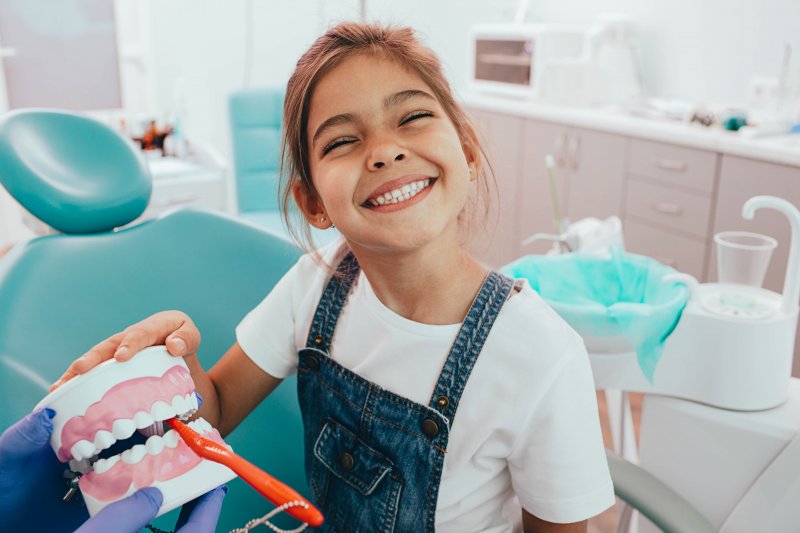 Girl in overalls smiling with teeth while holding toothbrush up to teeth model